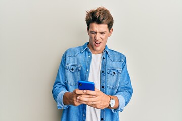 Handsome caucasian man using smartphone angry and mad screaming frustrated and furious, shouting...