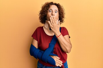 Beautiful middle age mature woman wearing cervical collar and arm on sling covering mouth with...