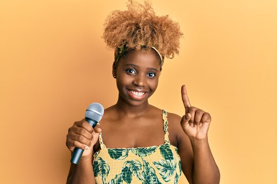 Young african woman with afro hair singing song using microphone smiling with an idea or question pointing finger with happy face, number one