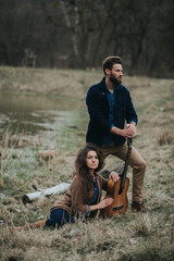 Naklejka na ściany i meble two caucasian lovers musicians with guitar standing by the lake. Young couple is hugging on autumn day outdoors. A bearded man and curly woman in love. Valentine's Day. Concept of love and family.