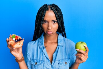 Beautiful hispanic woman holding pastry and healthy green apple depressed and worry for distress,...