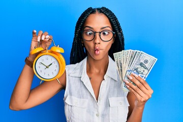 Beautiful hispanic woman holding alarm clock and dollars making fish face with mouth and squinting eyes, crazy and comical.