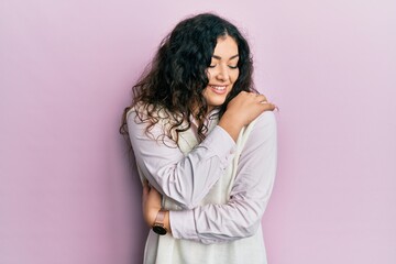 Young brunette woman with curly hair wearing casual clothes hugging oneself happy and positive,...