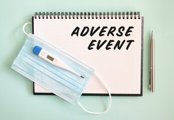 A medical protective mask and a thermometer are on the notepad. ADVERSE EVENT text in a notebook.