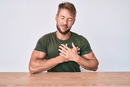 Young caucasian man wearing casual clothes sitting on the table smiling with hands on chest with closed eyes and grateful gesture on face. health concept.