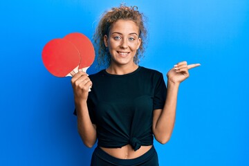 Beautiful caucasian teenager girl holding red ping pong rackets smiling happy pointing with hand...