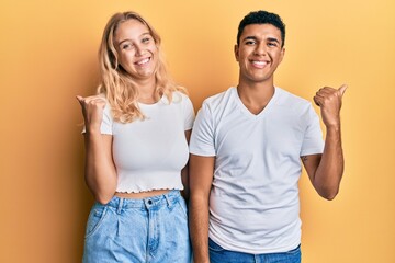 Young interracial couple wearing casual white tshirt smiling with happy face looking and pointing to the side with thumb up.