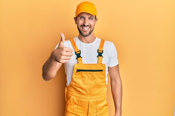 Young handsome man wearing handyman uniform over yellow background doing happy thumbs up gesture...