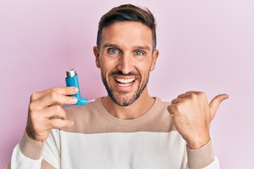 Handsome man with beard holding medical asthma inhaler pointing thumb up to the side smiling happy with open mouth - obrazy, fototapety, plakaty
