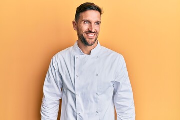 Handsome man with beard wearing professional cook uniform looking away to side with smile on face, natural expression. laughing confident.