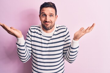 Naklejka premium Young handsome man wearing casual striped sweater standing over isolated pink background clueless and confused with open arms, no idea and doubtful face.