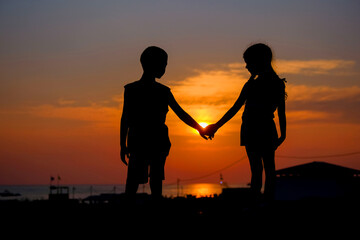 Fototapeta na wymiar Children hold hands in the background of the sunset. Silhouettes of a young boy and girl.