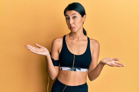 Beautiful asian young sport woman wearing sportswear and earphones clueless and confused expression with arms and hands raised. doubt concept.