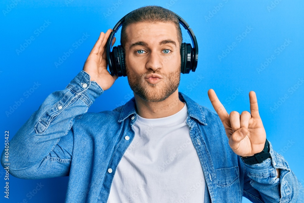 Wall mural Young caucasian man using headphones and doing rock symbol looking at the camera blowing a kiss being lovely and sexy. love expression.