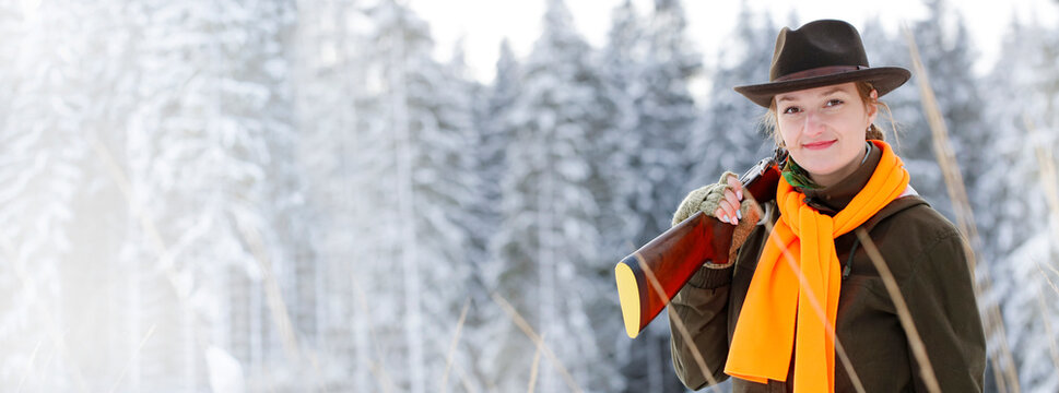 A young beautiful hunter woman on hunt in forest with rifle on the shoulder. Winter season. In the background are snowy trees. Banner photo.