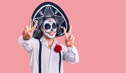 Man wearing day of the dead costume over background smiling with tongue out showing fingers of both hands doing victory sign. number two.