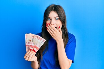 Young beautiful caucasian girl holding 10 colombian pesos banknotes covering mouth with hand, shocked and afraid for mistake. surprised expression