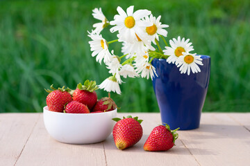 White bowl with sweet red strawberries and blue vase with chamomile flowers on green blurred background. Summer composition with seasonal berries
