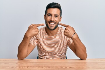Handsome hispanic man wearing casual clothes sitting on the table smiling cheerful showing and...