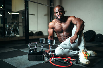Fototapeta na wymiar A man with a naked torso in gray pants sits on the floor next to dumbbells and a kettlebell. Strength training using sports equipment
