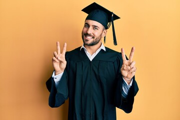 Young hispanic man wearing graduation cap and ceremony robe smiling looking to the camera showing...