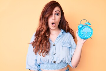 Young beautiful woman holding alarm clock scared and amazed with open mouth for surprise, disbelief face