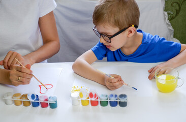A young mother and his cute little son in glasses draw together with paints on white paper.