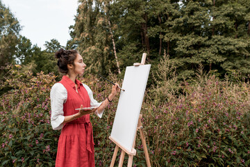 Beautiful woman in red dress white shirt, in summer park forest, draws a picture, white canvas paint and a brush, free space for a copy of text. Art creativity concept.