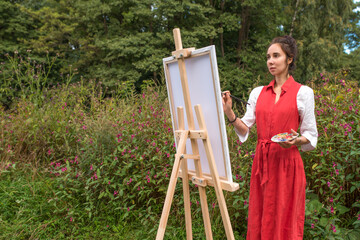 Beautiful woman in a red dress white shirt, summer park forest, draws picture, creating landscape free space copy text. Creativity and art concept. Trees and bushes background.
