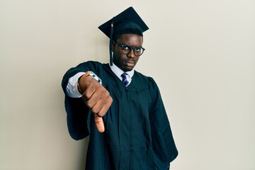 Handsome black man wearing graduation cap and ceremony robe looking unhappy and angry showing rejection and negative with thumbs down gesture. bad expression.