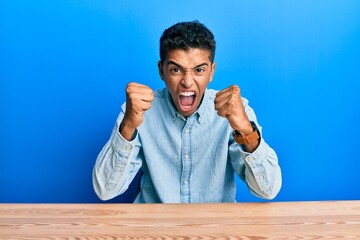 Young handsome african american man wearing casual clothes sitting on the table angry and mad...