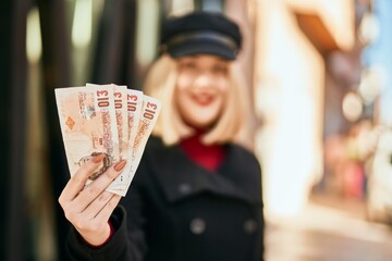 Young blonde woman smiling happy holding united kingdom pounds banknotes at the city.