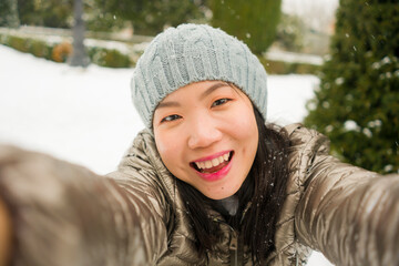 winter lifestyle portrait of young happy and beautiful Asian Chinese woman taking selfie picture with mobile phone enjoying snow at city park during Christmas holidays travel