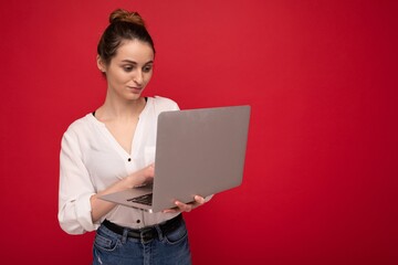 Side profile of Beautiful brunet young woman holding netbook computer looking down wearing white shirt typing on keyboard isolated on red background