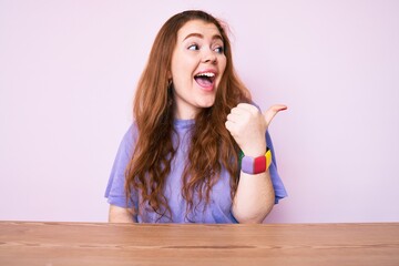 Young redhead woman wearing casual clothes sitting on the table pointing thumb up to the side smiling happy with open mouth