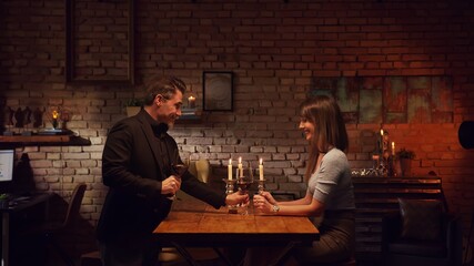 Elegant couple having romantic dinner at home, sitting at table in living room drinking red wine, talking. Stay at home concept.