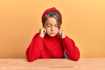 Little beautiful girl wearing casual clothes sitting on the table covering ears with fingers with annoyed expression for the noise of loud music. deaf concept.