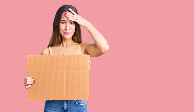 Beautiful brunette young woman holding cardboard banner with blank space stressed and frustrated with hand on head, surprised and angry face