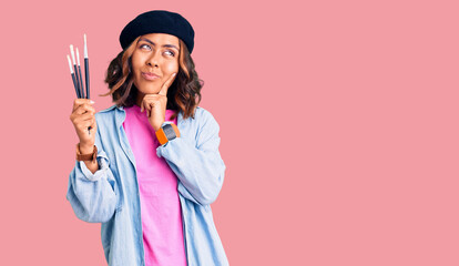 Young beautiful mixed race woman holding paintbrushes serious face thinking about question with...