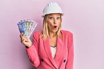 Beautiful middle age blonde woman wearing architect hardhat holding indian rupee scared and amazed with open mouth for surprise, disbelief face