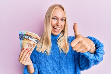 Young blonde girl holding canadian dollars approving doing positive gesture with hand, thumbs up smiling and happy for success. winner gesture.