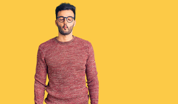 Young handsome hispanic man wearing winter sweater and glasses making fish face with lips, crazy and comical gesture. funny expression.
