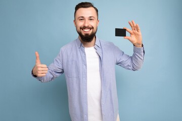 photo shot of Handsome smiling brunette bearded young man wearing stylish blue shirt and white t-shirt isolated over blue background wall holding credit card looking at camera and showing thumbs up