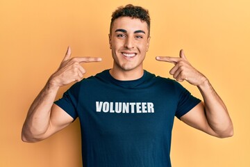 Hispanic young man wearing volunteer t shirt smiling cheerful showing and pointing with fingers teeth and mouth. dental health concept.