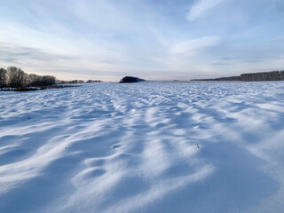 Obraz premium A snow-covered field under a blue sky with feathery clouds. Winter landscape in blue tones with a snow relief surface with trees in the background.