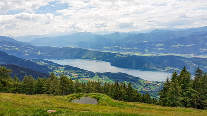 Fototapeta na wymiar A panoramic view on the Millstaettersee lake from Granattor in Austrian Alps. The distant lake is surrounded by high mountains. Few clouds above. Endless mountain chains. Lush and vast pasture. Relax
