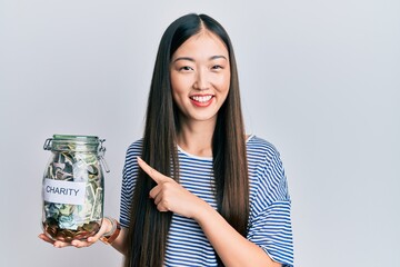 Young chinese woman holding charity jar with money smiling happy pointing with hand and finger