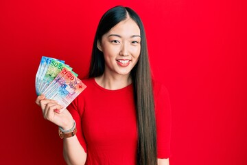 Young chinese woman holding swiss franc banknotes looking positive and happy standing and smiling with a confident smile showing teeth