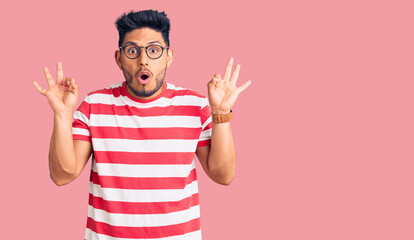 Fototapeta premium Handsome latin american young man wearing casual clothes and glasses looking surprised and shocked doing ok approval symbol with fingers. crazy expression