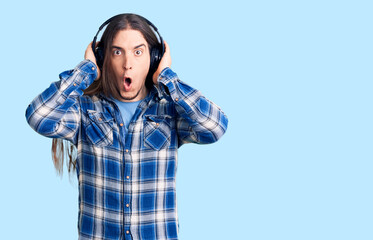 Young adult man with long hair listening to music using headphones scared and amazed with open mouth for surprise, disbelief face